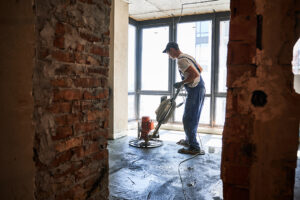 Male worker using troweling machine while screeding floor in apartment under renovation. Man finishing concrete surface with floor screed grinder machine in room with large window.