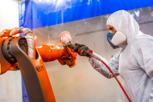 Painter with protective gear using a spray gun to paint an industrial robot arm in a factory