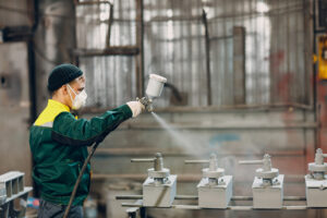 Powder primer coating of metal parts. Worker man in a protective suit sprays powder paint from gun on metal product construction at factory plant.