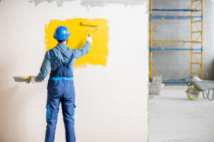 Workman in uniform painting wall with yellow paint at the construction site indoors